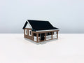 Brown log-style miniature cabin with a black roof, white porch railing and gray steps, shown from an angle.